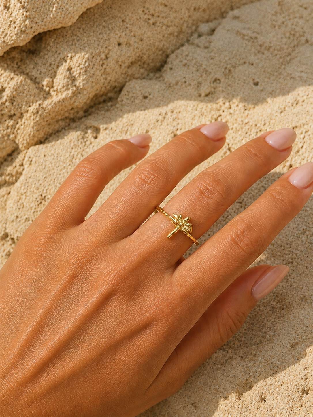 A close-up of a hand resting on sandy ground, wearing a delicate gold ring with a small cross and floral design on the ring finger.
