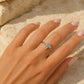 A silver ring with a small dragonfly design set with clear stones, worn on a hand with pale pink manicured nails, resting on a sandy beige background.