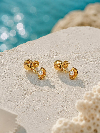 A pair of gold shell stud earrings with small white pearl accents, displayed on a textured white stone surface with a blurred turquoise ocean background.