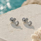 A pair of silver seashell stud earrings with small pearl accents, displayed on a textured white stone surface with a blurred turquoise ocean background.