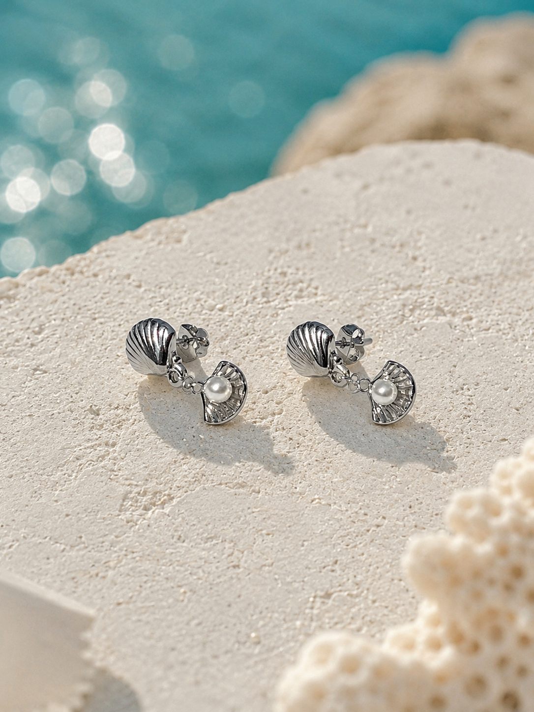 A pair of silver seashell stud earrings with small pearl accents, displayed on a textured white stone surface with a blurred turquoise ocean background.