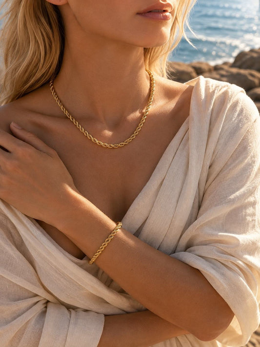 A close-up lifestyle shot of a woman wearing a gold rope chain necklace and matching gold rope bracelet, styled with a cream-colored linen wrap top against a sunny beach background.