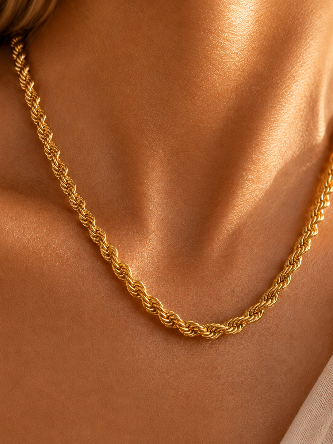A close-up of a gold rope chain necklace worn on a model's neck, showing the detailed twisted links of the chain against warm skin tones.