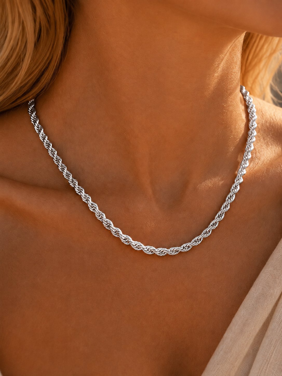 A close-up of a silver twisted rope chain necklace worn on a model's neck, with warm golden sunlight highlighting the metallic texture.