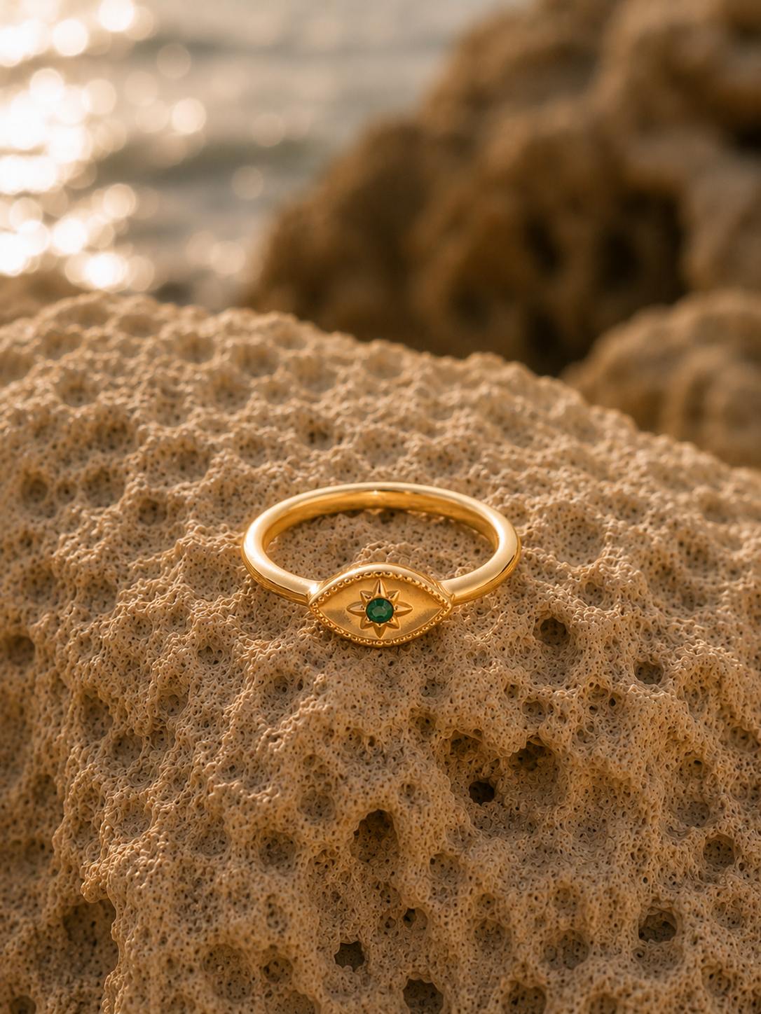 A gold eye-of-providence ring with a green gemstone, resting on a textured sandy rock with a blurred beach background.