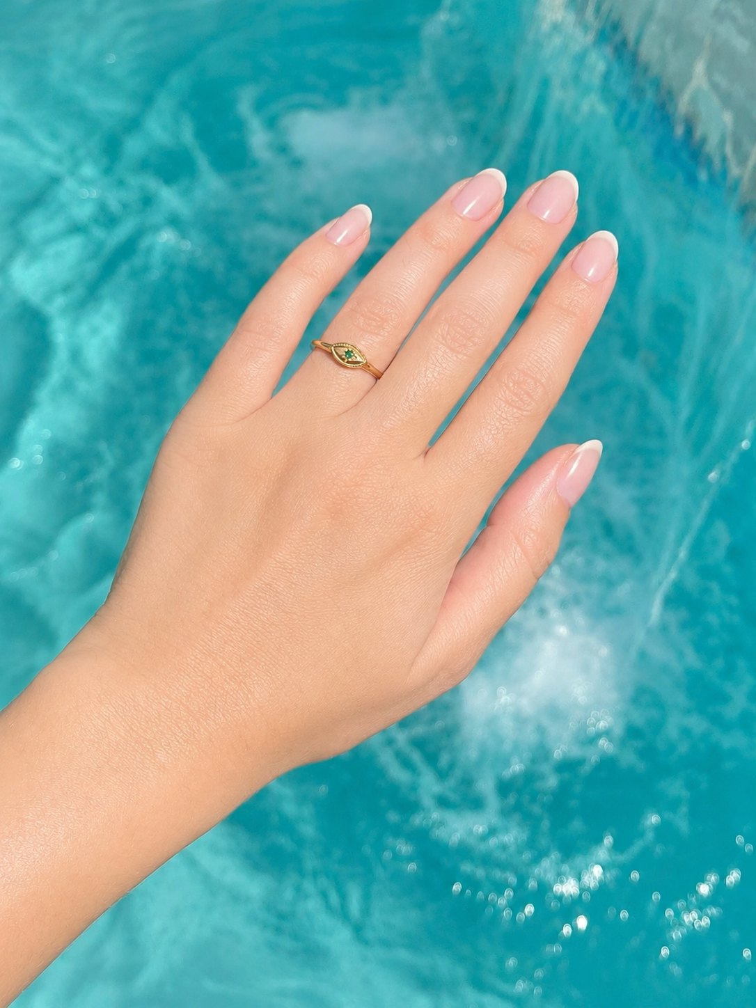 A gold ring with a small green stone set on a person's ring finger, shown against a bright turquoise swimming pool background.