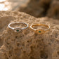 Two eye-shaped rings, one in silver and one in gold, each featuring a small green gemstone, displayed on a textured sandy rock with a blurred beach background.