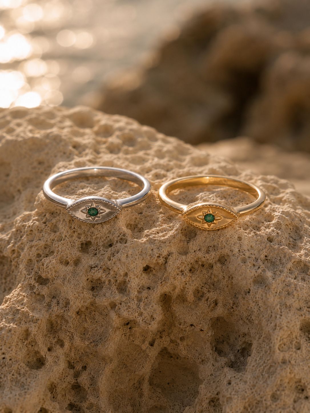 Two eye-shaped rings, one in silver and one in gold, each featuring a small green gemstone, displayed on a textured sandy rock with a blurred beach background.