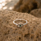 A silver eye-shaped ring with a green center stone and small clear stones, resting on a textured sandy rock with a blurred beach background.