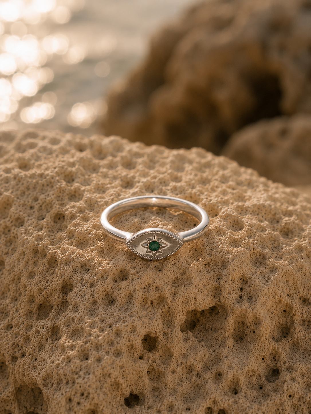 A silver eye-shaped ring with a green center stone and small clear stones, resting on a textured sandy rock with a blurred beach background.