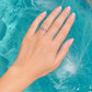 A silver-toned ring with a small green stone set on a woman's ring finger, shown with a French manicure against a bright turquoise swimming pool background.
