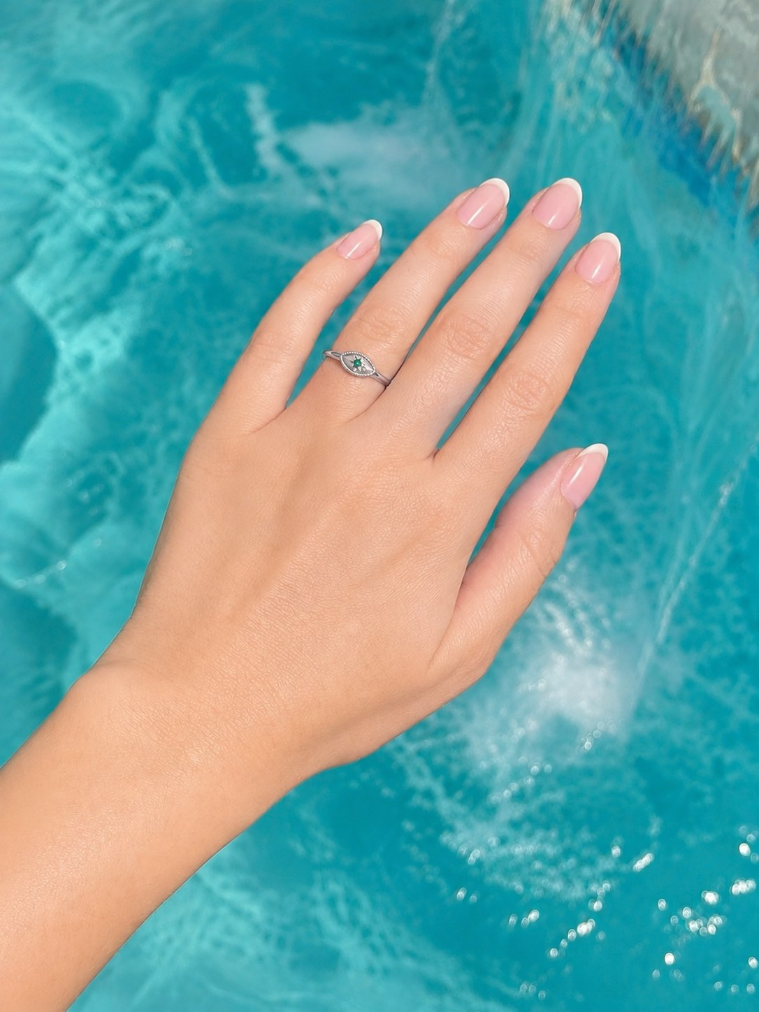 A silver-toned ring with a small green stone set on a woman's ring finger, shown with a French manicure against a bright turquoise swimming pool background.