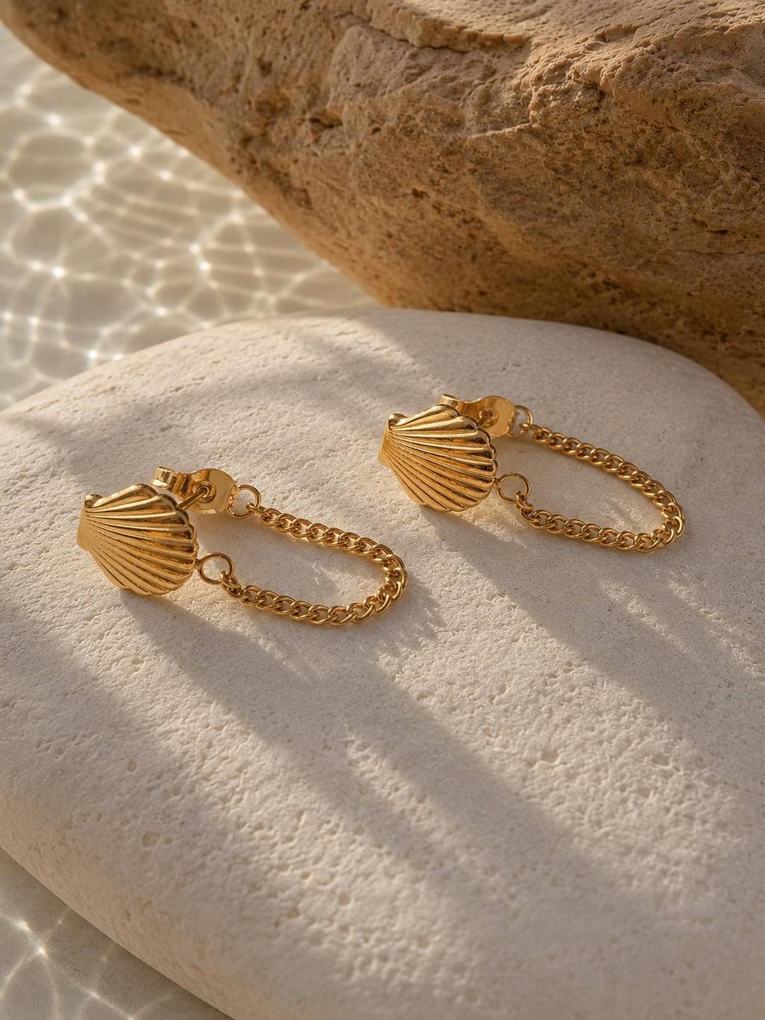 A pair of gold shell-shaped dangle earrings with chain links, displayed on a textured white stone with a sandy beach and rock background under warm sunlight.