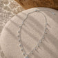 A silver-toned necklace with a small shell-shaped pendant and dangling bead accents, arranged on a round stone surface with warm sunlight and shadows.