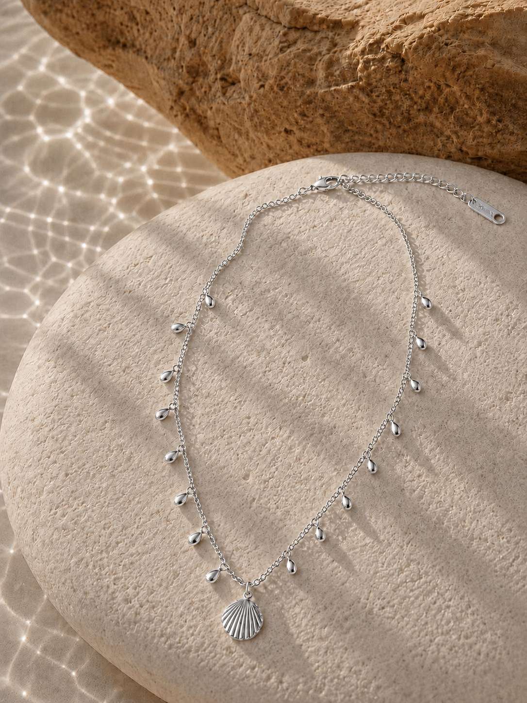 A silver-toned necklace with a small shell-shaped pendant and dangling bead accents, arranged on a round stone surface with warm sunlight and shadows.