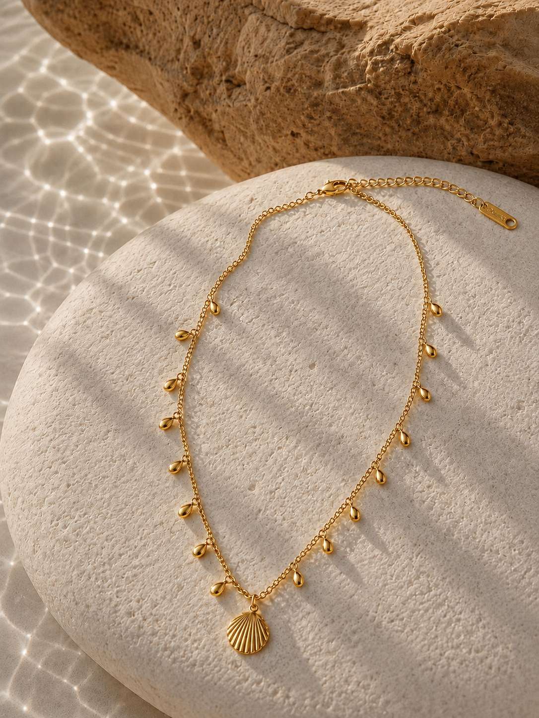 A gold-toned necklace with a small shell-shaped pendant and dangling bead accents, arranged on a round stone with a larger rock in the background and sunlight filtering through water ripples.