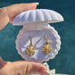 A close-up of a hand holding an open white clamshell jewelry box containing a pair of gold dangle star earrings with textured circular pendants and small clear stones above them, against a blurred blue water background.