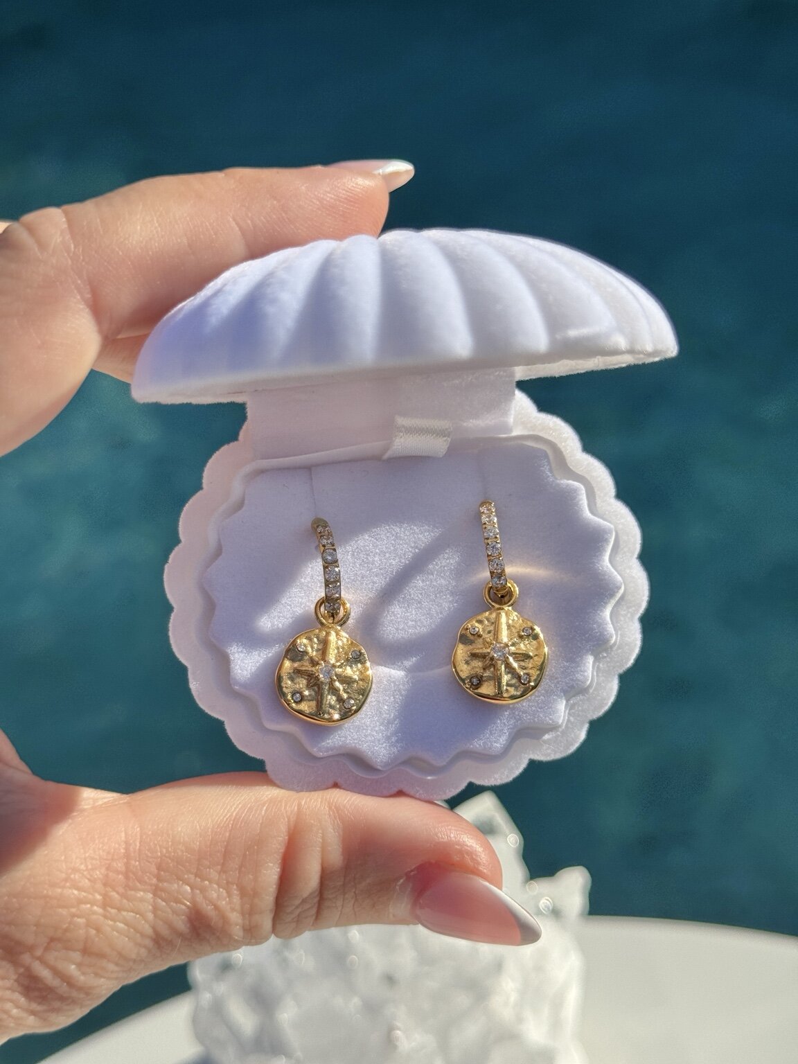 A close-up of a hand holding an open white clamshell jewelry box containing a pair of gold dangle star earrings with textured circular pendants and small clear stones above them, against a blurred blue water background.