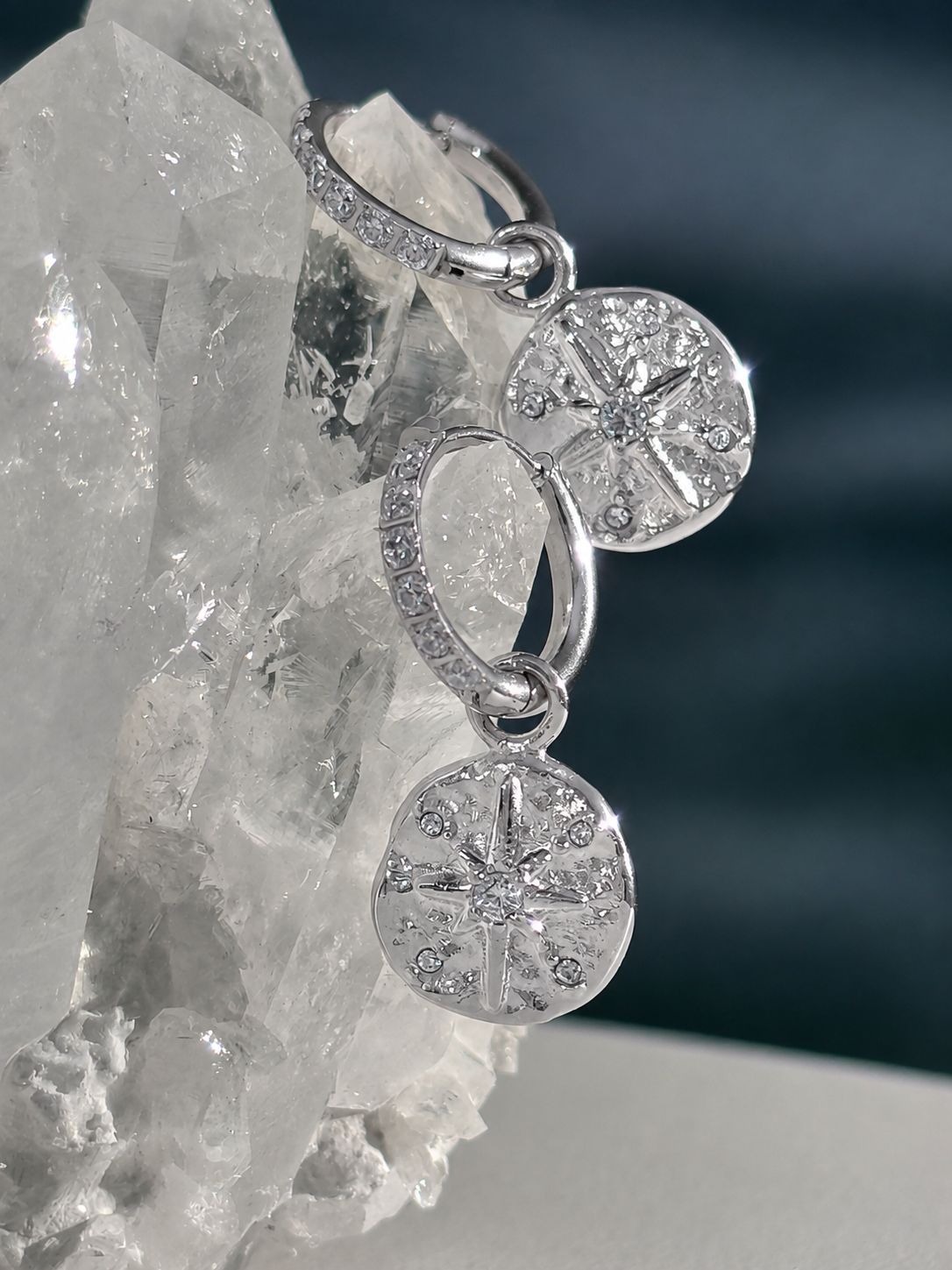 Sterling silver drop earrings with circular starburst charms and pavé-set stones, displayed on a clear crystal cluster against a dark blue background.