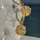 A pair of gold hoop earrings with dangling round starburst charms, each set with small clear stones, displayed against a clear crystal cluster and dark blue background. Waterproof Earrings Jewelry