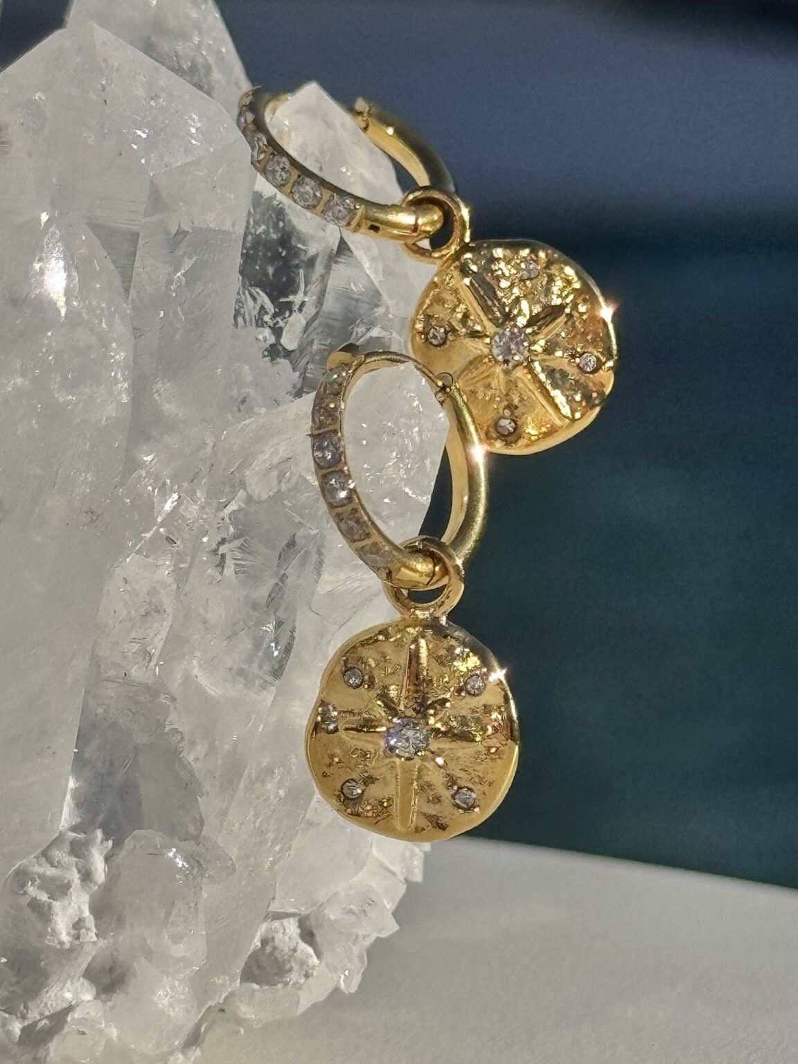 A pair of gold hoop earrings with dangling round starburst charms, each set with small clear stones, displayed against a clear crystal cluster and dark blue background. Waterproof Earrings Jewelry