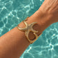 A gold-toned starfish-shaped cuff bracelet worn on a person's wrist, with a bright turquoise swimming pool background.
