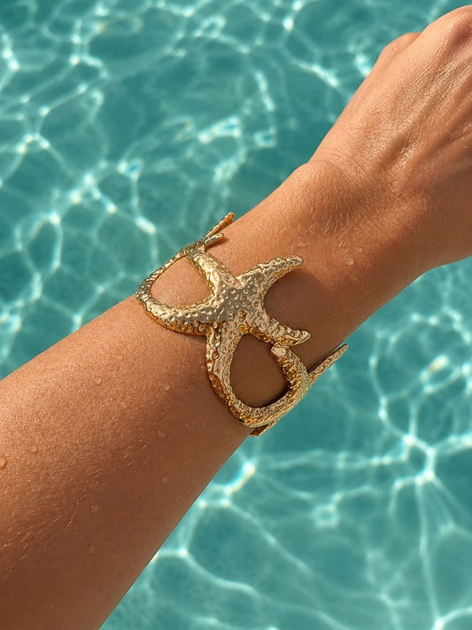 A gold-toned starfish-shaped cuff bracelet worn on a person's wrist, with a bright turquoise swimming pool background.