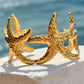 A textured gold cuff bracelet designed like a starfish, resting on a white stone ledge with a blurred blue ocean background.