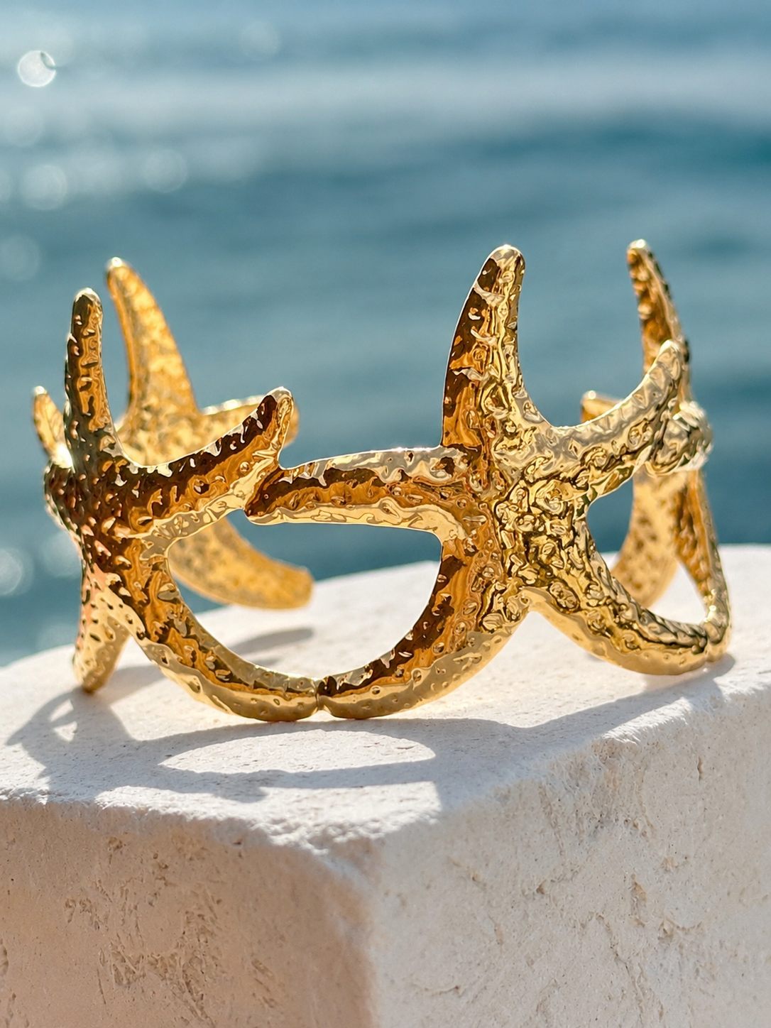 A textured gold cuff bracelet designed like a starfish, resting on a white stone ledge with a blurred blue ocean background.