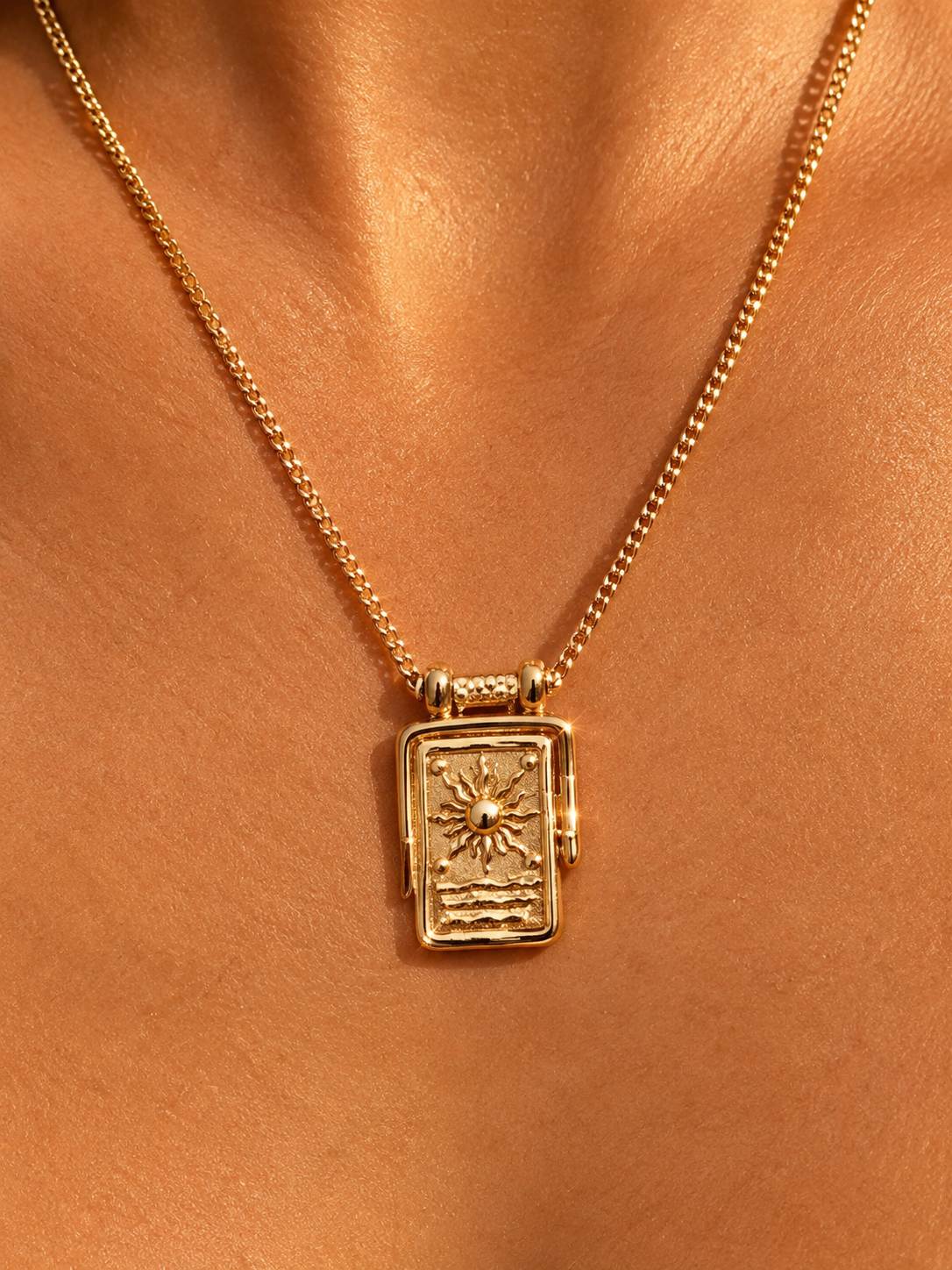 A gold chain necklace with a rectangular pendant featuring a sun and wave design, shown worn on a model's neck against a warm skin-toned background.
