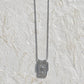 A silver-toned necklace with a rectangular pendant featuring a sun and palm tree motif, hanging against a textured white stucco wall with a turquoise blue background at the top.