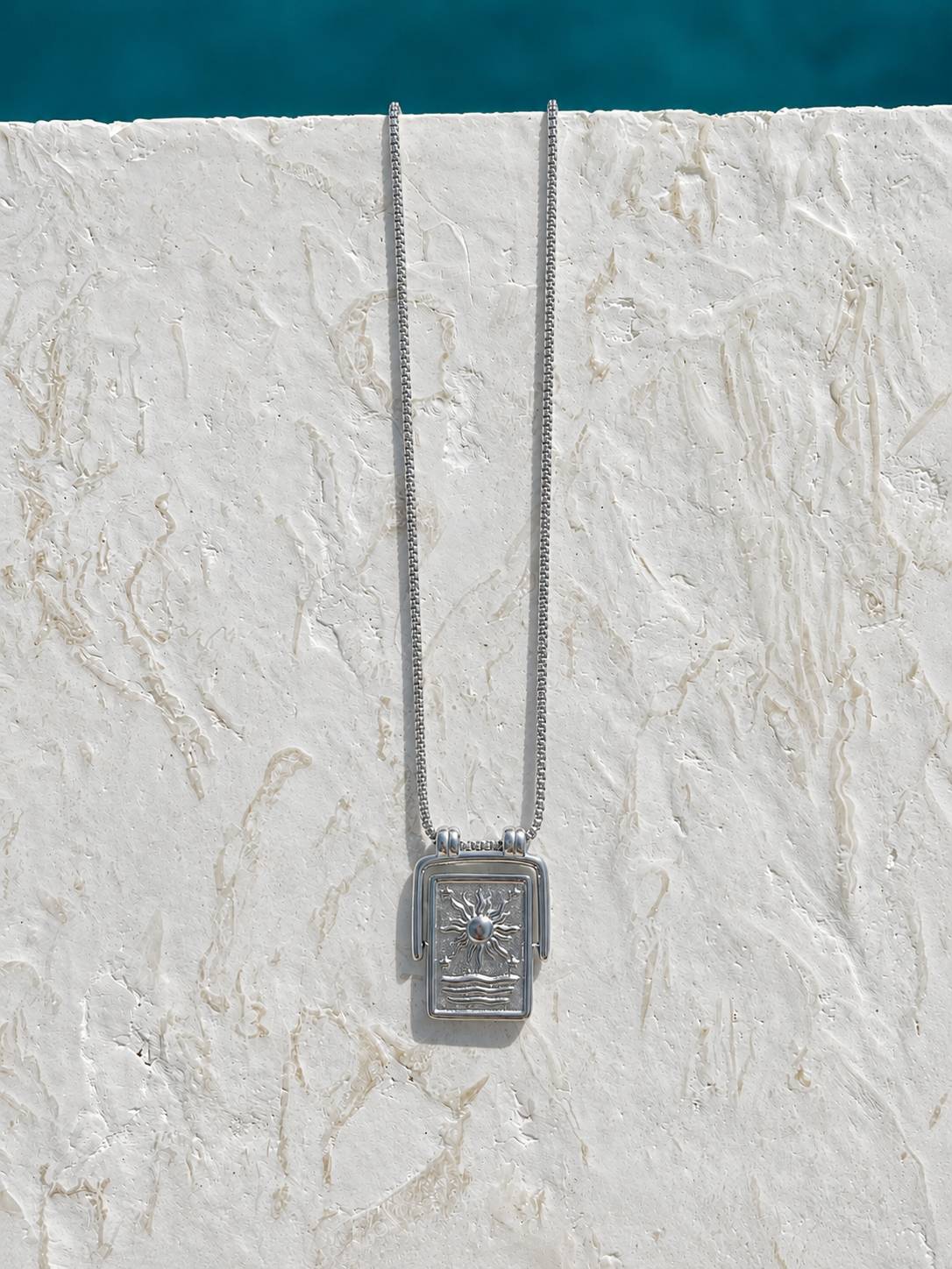 A silver-toned necklace with a rectangular pendant featuring a sun and palm tree motif, hanging against a textured white stucco wall with a turquoise blue background at the top.