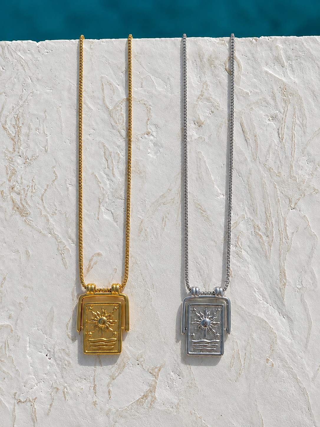 Pendant sun necklaces hanging from chains on a textured white stone surface. One necklace is gold-toned with a gold pendant, and the other is silver-toned with a silver pendant.