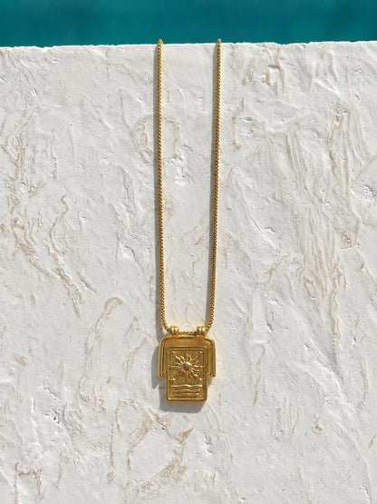 A gold necklace with a rectangular sun pendant featuring a stylized sun and geometric patterns, hanging against a textured white stucco wall with a turquoise pool edge visible at the top.
