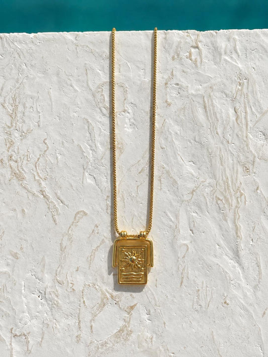 A gold necklace with a rectangular sun pendant featuring a stylized sun and geometric patterns, hanging against a textured white stucco wall with a turquoise pool edge visible at the top.