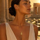 A woman with her eyes closed wears a delicate gold necklace with a square pendant, set against a warm, sunlit background of ancient stone columns and water lilies.