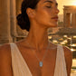A silver pendant necklace with a rectangular engraved design, set against a warm, golden background of ancient stone columns and water lilies.