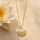 A gold lotus pendant necklace with a delicate chain, displayed on a textured beige stone surface next to a white lotus flower.