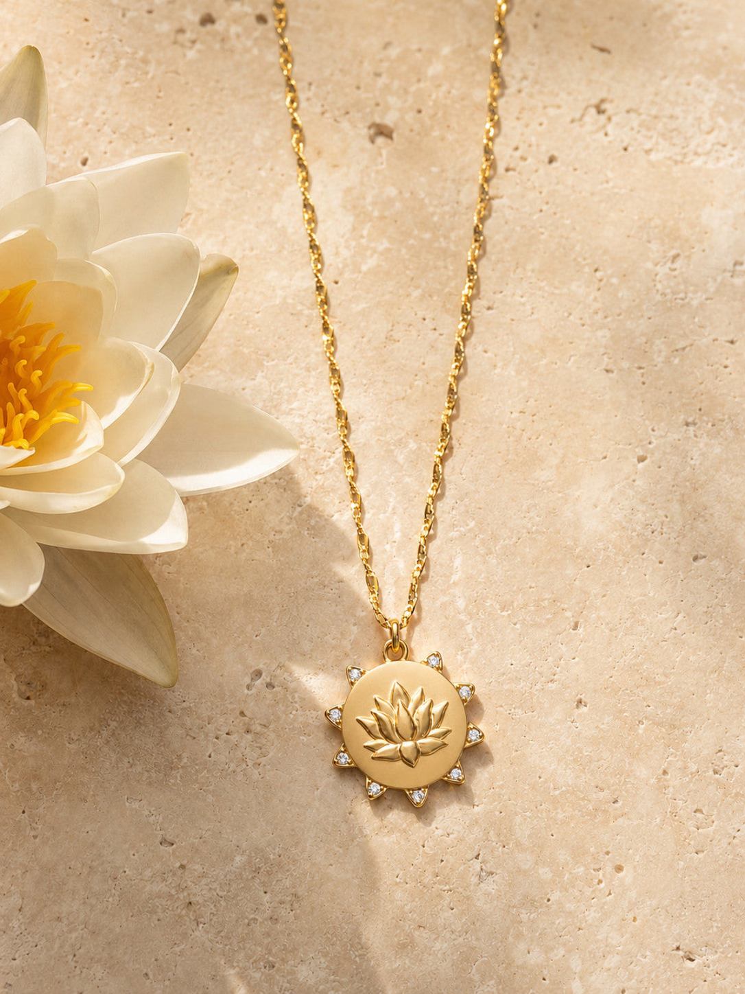 A gold lotus pendant necklace with a delicate chain, displayed on a textured beige stone surface next to a white lotus flower.