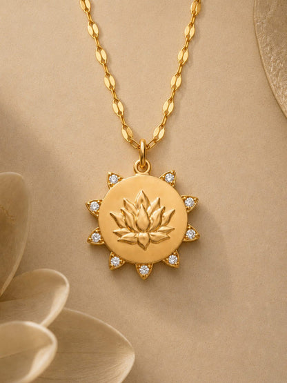 A gold pendant necklace featuring a lotus flower design in the center, surrounded by small clear stones, hanging from a gold chain against a warm beige background with soft decorative petals.
