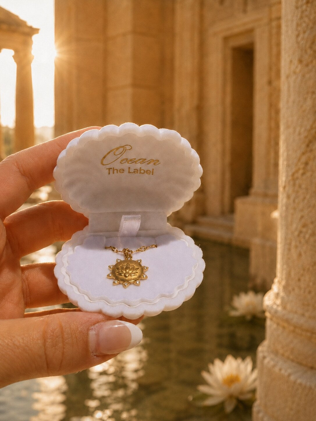 A hand holds an open white clamshell jewelry box with the brand name 'Ocean The Label' printed inside. The box contains a gold pendant necklace.