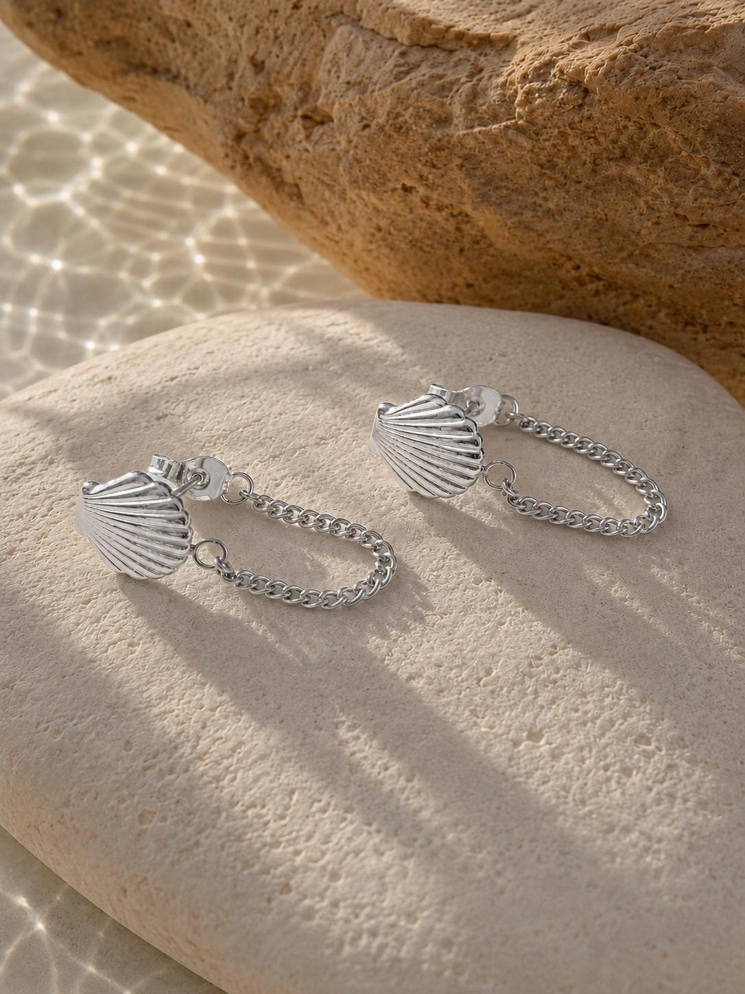 A pair of silver shell-shaped drop earrings with short chain links, displayed on a textured beige stone with a larger rock in the background.