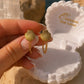 A hand holds a pair of gold shell-shaped dangle earrings in front of an open white seashell jewelry box branded 'Ocean The Label', with rocks and water in the background.