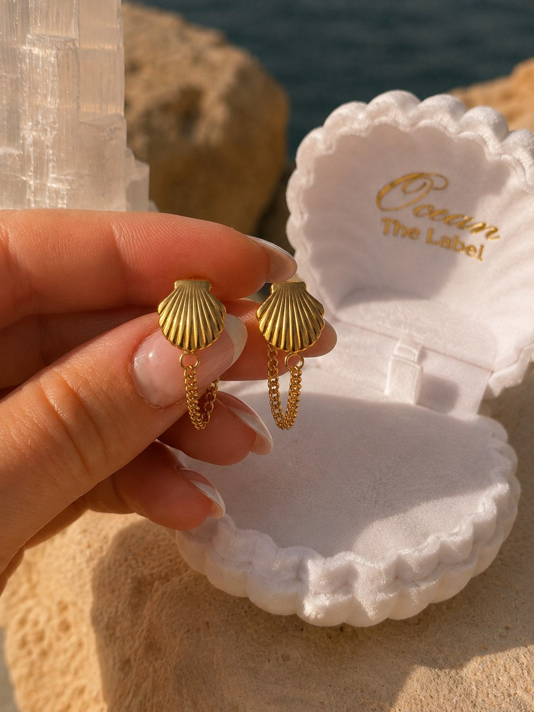 A hand holds a pair of gold shell-shaped dangle earrings in front of an open white seashell jewelry box branded 'Ocean The Label', with rocks and water in the background.