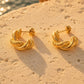 A pair of gold twisted hoop earrings resting on a textured beige stone surface with a blurred background of water and warm sunlight.