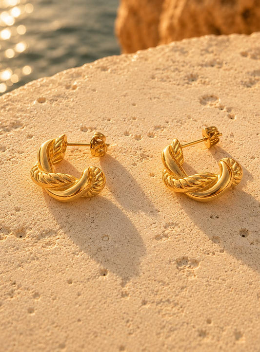 A pair of gold twisted hoop earrings resting on a textured beige stone surface with a blurred background of water and warm sunlight.