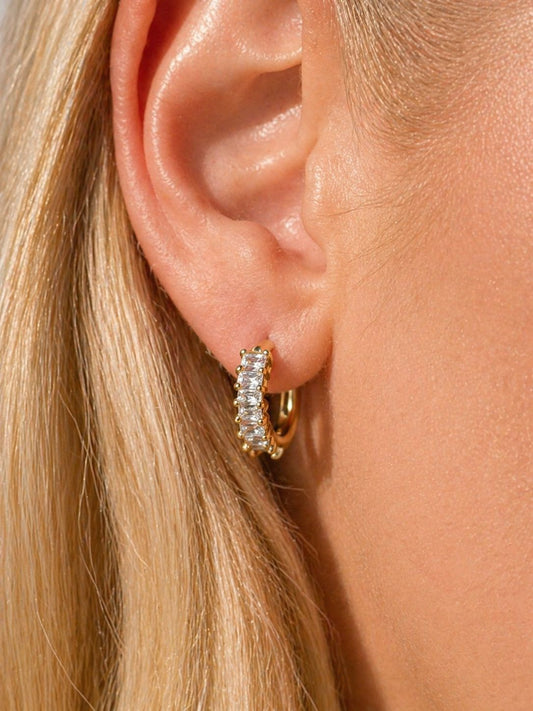 A close-up of a model's ear wearing a small gold huggie hoop earring set with clear pavé gemstones.