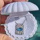 A silver rectangular pendant necklace with a bright blue oval zircon aquamarine gemstone, displayed inside a white clamshell jewelry box held by a hand against a teal background. Protection Necklace