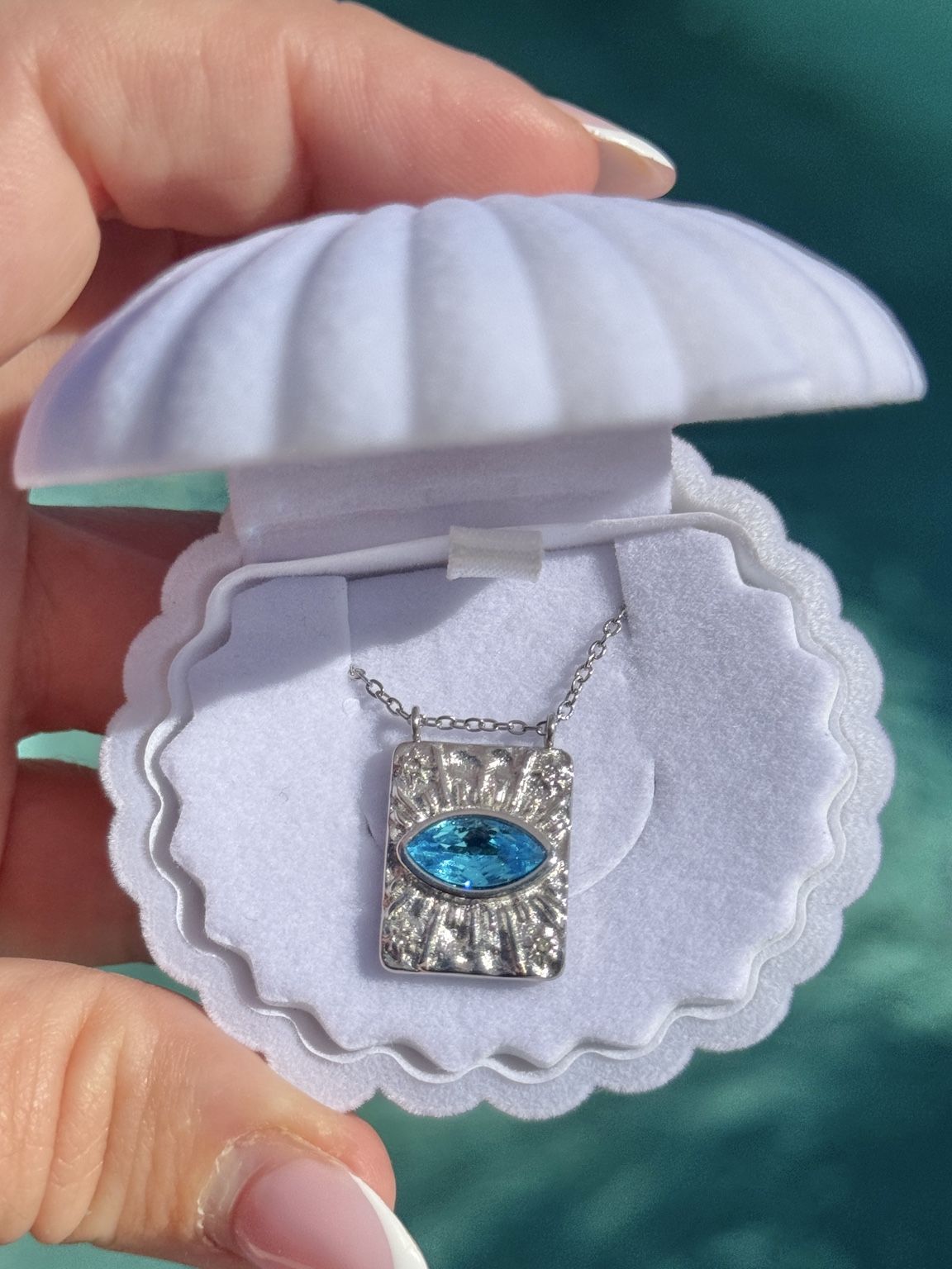 A silver rectangular pendant necklace with a bright blue oval zircon aquamarine gemstone, displayed inside a white clamshell jewelry box held by a hand against a teal background. Protection Necklace