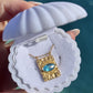 A gold pendant necklace with a marquise-cut light blue gemstone, displayed inside a white clamshell jewelry box held by a hand against a blurred teal background.